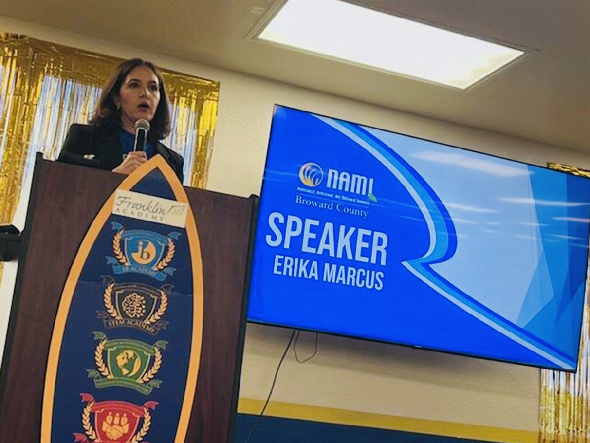 Erika Marcus speaking into a microphone at a podium, her name shown as speaker on the screen behind her